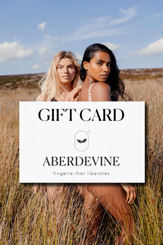 Two women standing in a field with an Aberdevine gift card overlay. The card is white with the words 'gift card', the Aberdevine feather logo and text as well as the tagline 'lingerie that liberates'