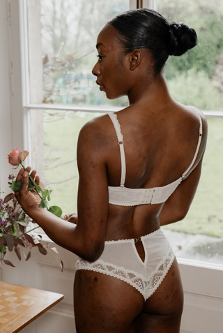 Woman wearing a white Aberdevine lace lingerie set holding flowers by a window.